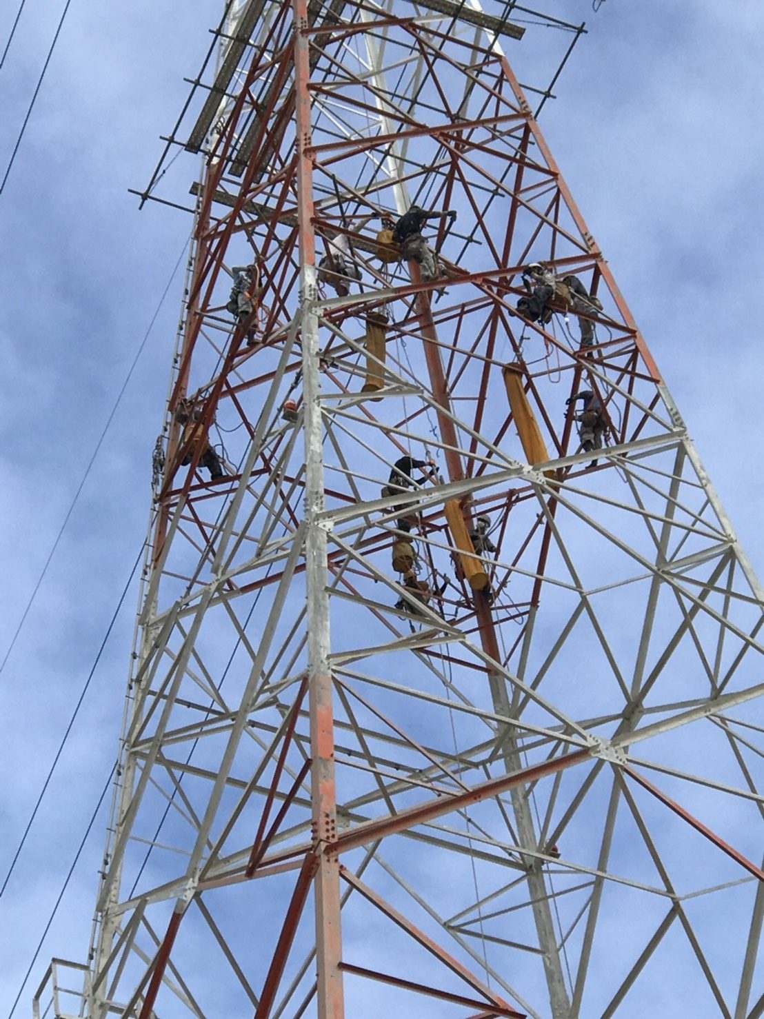 紅白の送電線鉄塔を見上げた風景。鉄骨の各所で塗装作業を行う職人たち